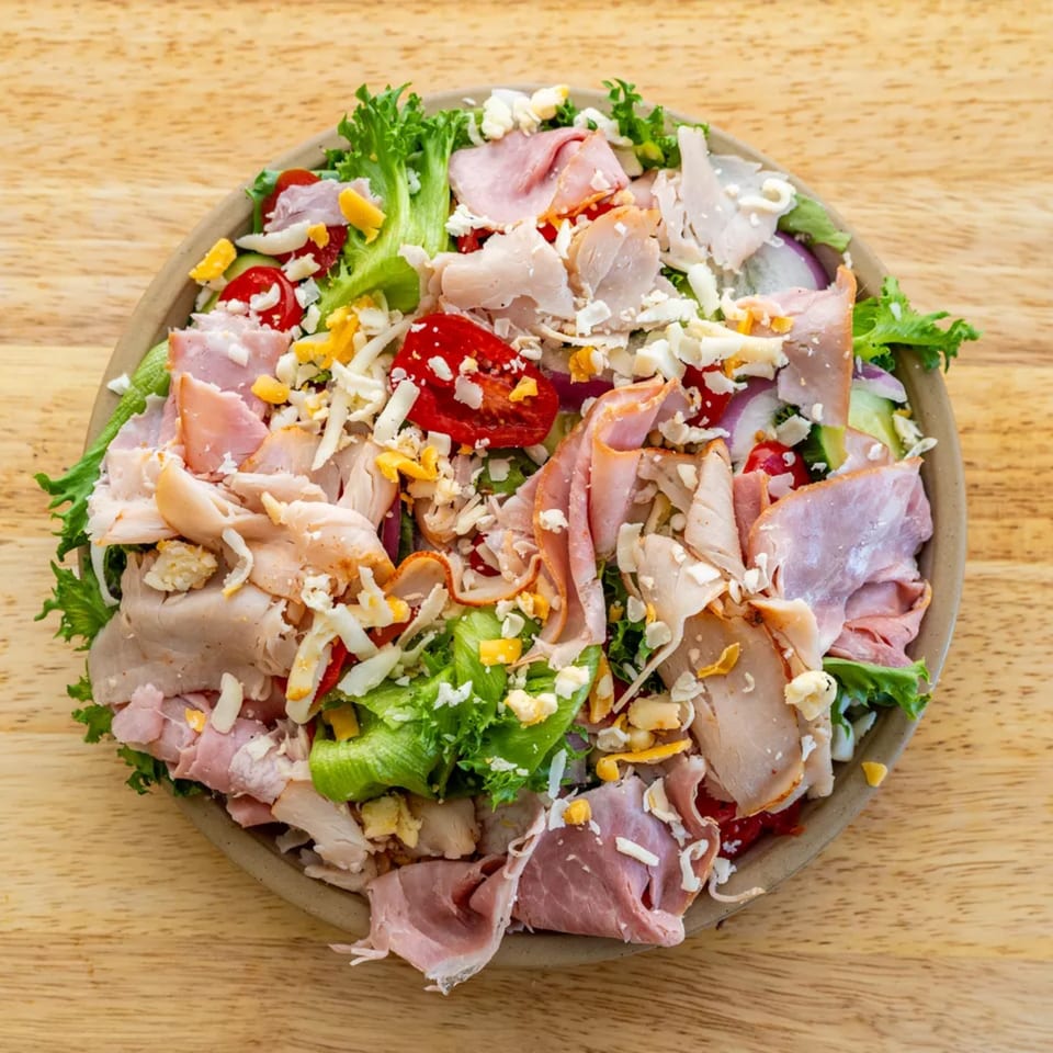 Overhead view of a chef salad with ham, turkey, cheese, tomatoes, and lettuce on a wooden table.